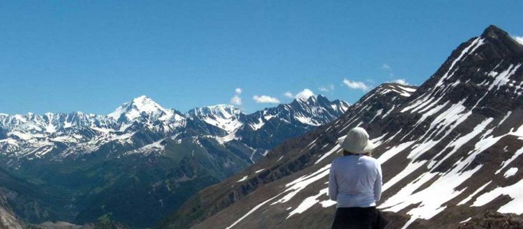 A woman stands off to the right hand side of the picture. Her back is to the camera. She is wearing a long white sleeved shirt and a white bucket hat. She is looking out toward numerous snow capped mountain peaks. This is the Alps.
