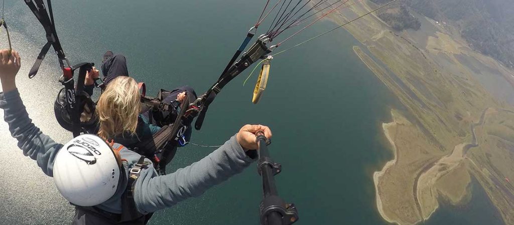 Blond woman is paragliding. You cannot see her face. The pilot is sitting behind her. They are overlooking a vast body of blueish-green water and land in the distance.
