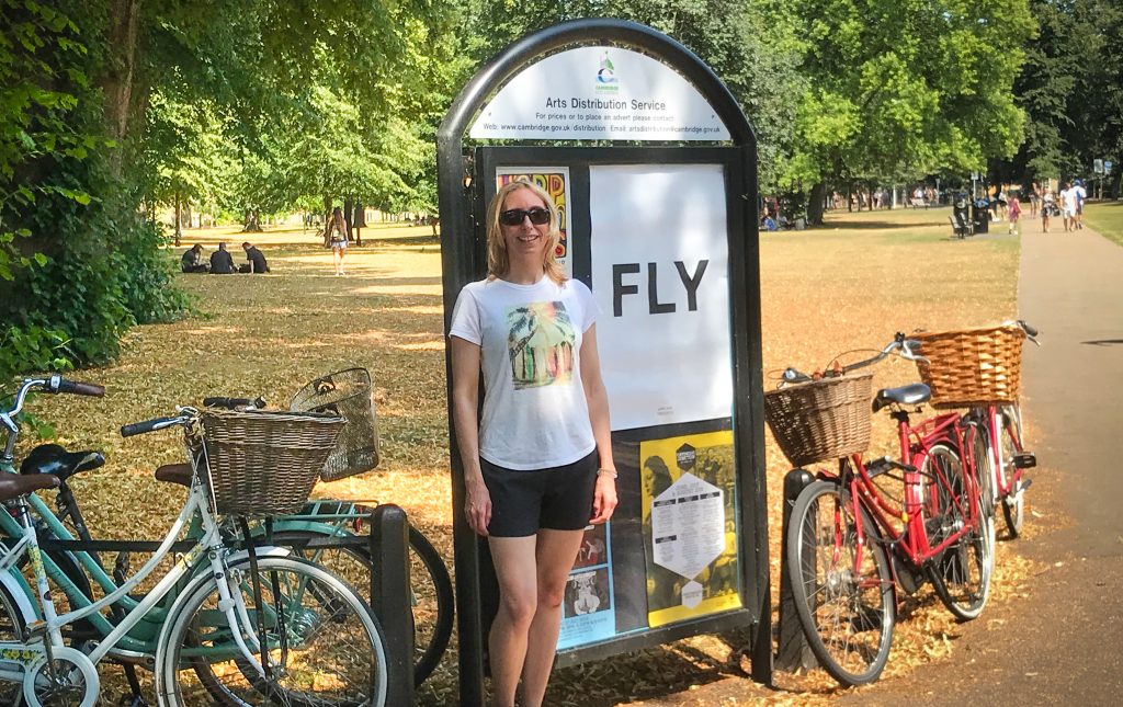 Melissa Rodway - standing in front of a sign that says FLY. It is a summer day, and there are bikes on either side of Melissa and the sign.